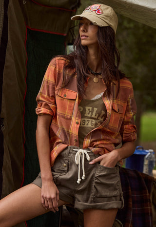 a-woman-wearing-a-tan-baseball-cap-an-orange-and-brown-plaid-flannel-shirt-a-brown-graphic-tank-top-and-olive-cargo-shorts-posed-in-front-of-a-tent-in-an-outdoor-setting