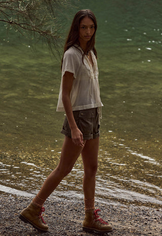 a-woman-walking-along-a-rocky-shoreline-next-to-a-body-of-water-wearing-a-loose-white-short-sleeve-top-dark-gray-shorts-and-tan-hiking-boots-with-red-laces